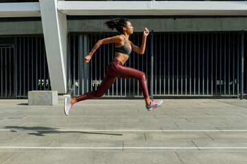 Strong sportswoman in comfortable tracksuit runs jumping up past modern building on sunny city street side view