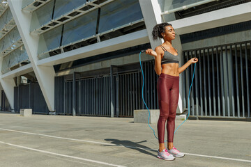 Concentrated young  woman in stylish tracksuit jumps rope near modern building on sunny city street
