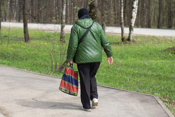 An elderly woman in casual clothes with a bag walks down the street in the park. View from the...