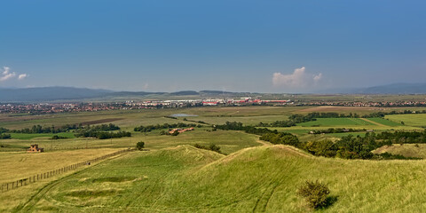 Transylvanian landscape with green fields with and a city and mountains in the distance on a sunny day with clear blue sky, view from above ffrom Rapa Rosie, Romania 