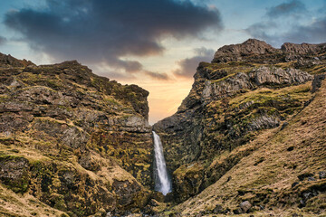 Waterfall flowing down the rocks covered with moss under a gray cloudy sky during the sunset