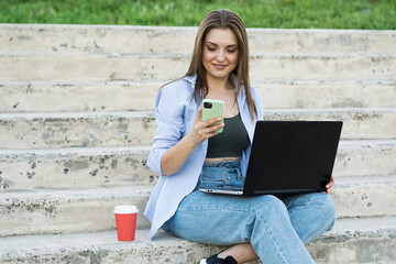 Happy and attractive caucasian girl sitting with a laptop on the steps and using a smartphone. Distance working. Freelance concept