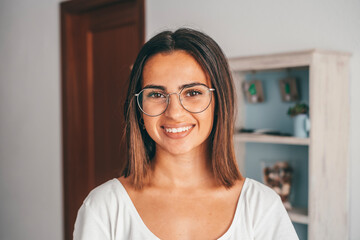 Happy young woman in eyeglasses looking at camera from domestic room of her house. Beautiful satisfied lady with shrt hair smiling while facing camera at modern apartment. Female youth smiling at home