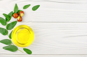 Jojoba oil in a bowl, a sprig of jojoba and fruits on a white wooden table
