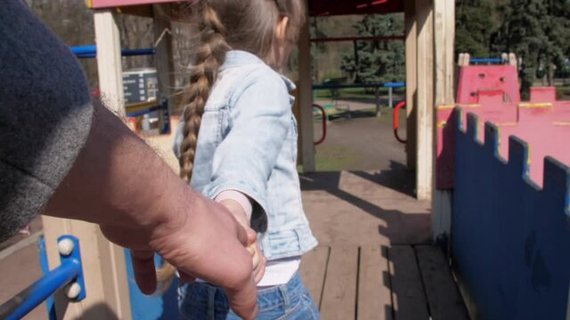 Blonde young schoolgirl holds stepfather hand and walks down brown wooden attraction stairs closeup