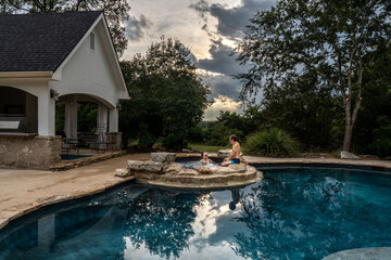 A mature man and woman enjoy a dip in a spa connected, above, a swimming pool with a cabana and a waterfall, Hill Country, Texas