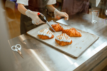 Woman confectioner burns albumenous cream decorating croissant at metal countertop