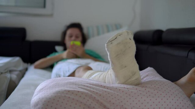 Young woman with foot bone fracture resting on couch at home and surfing on her smartphone after a foot injury accident