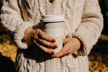 Closeup of a woman wearing a knitted sweater, holding a reusable coffee cup outdoors.