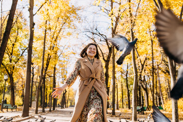 Young woman running among pigeons in a city central park on warm autumn day.