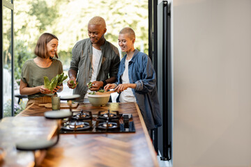 Friends cooking salad at home kitchen. Black man and european girls enjoying time together. Concept of healthy eating. Modern lifestyle friendship. Interior of apartment. Sunny daytime