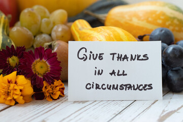 A handwritten quote to give thanks in all circumstances with pumpkin, grapes, and fall flowers. A...
