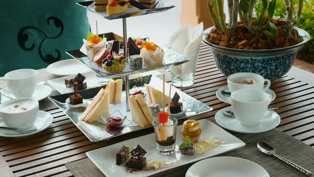 Traditional English afternoon tea stand with selection of sweets - cakes, snacks, sandwiches and macaroons. Tea set and coffee with assortment of pastries in outdoor cafe. Sweet buffet breakfast
