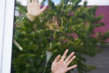 Young woman in black medical mask looking out window, putting hands on window being isolated at home during coronavirus epidemic infection. Thoughtful female with brown eyes observing through window.