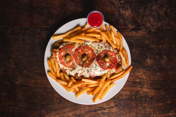 Overhead view of a Neapolitan-style milanesa with french fries on a plate on a wooden table.