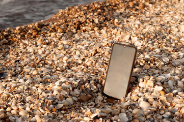 A black cell phone stands on a seashell beach next to the sea in the setting sun