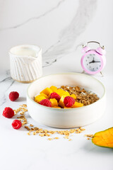 Bowl of granola with yogurt, milk and fresh raspberries, blueberries, mango for a healthy breakfast on a white marble table
