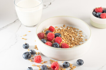 Bowl of granola with yogurt, milk and fresh raspberries, blueberries, mango for a healthy breakfast on a white marble table