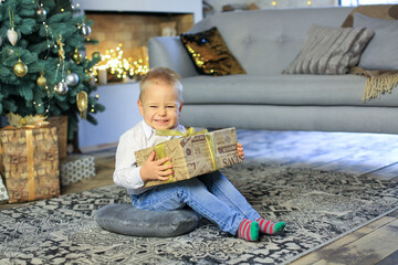 Little happy boy holding a box with a new year gift sits on the floor against the background of a christmas tree and fireplace