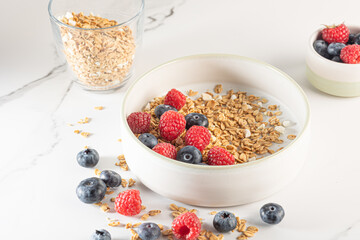 Bowl of granola with yogurt, milk and fresh raspberries, blueberries, mango for a healthy breakfast on a white marble table
