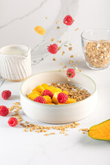 Bowl of granola with yogurt, milk and fresh raspberries, blueberries, mango for a healthy breakfast on a white marble table
