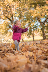 Happy toddler girl in pink coat jacket joyfully walking through autumn leaves in park setting