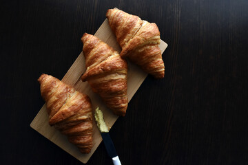 Croissants and a knife with butter on a bamboo chopping board on dark brown wood table. 