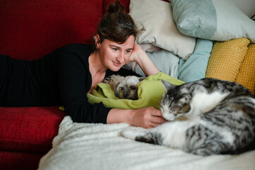 Beautiful woman in late 30s lying on couch with a dog and a cat. Caucasian female playing at home with her pets.