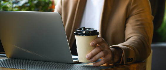 Cropped view of businessman in coat holding takeaway coffee near laptop in cafe, panorama 