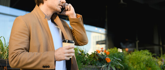 Cropped view of good looking businessman in coat holding coffee to go and talking on cellphone on urban street, panorama 