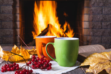 Mugs of tea or coffee, autumn leaves and viburnum berries before cozy fireplace, in country house, autumn or winter holidays.