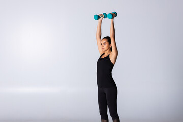 Woman lifting dumbbells over head on white background in black sportswear at gym, trainer show exercise with weights muscle building