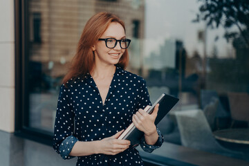 Businesswoman with red hair carries modern device and notepad goes on formal meeting