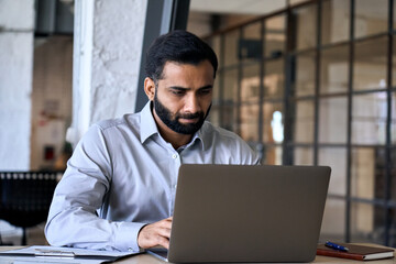 Handsome serious concentrated focused young Indian Hispanic ceo businessman using computer working in modern office, having virtual training on laptop typing watching online webinar. - Powered by Adobe