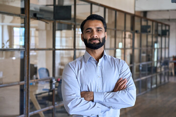 Confident happy successful ceo financial manager. Bearded indian businessman looking at camera standing in modern office with arms crossed. Handsome classy corporation owner. Business portrait.