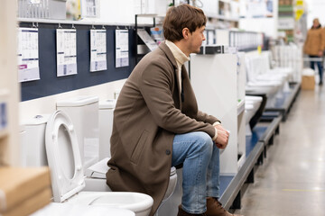 European man in a brown coat is sitting on the toilet in a hardware store, choosing a toilet for the bathroom during repairs