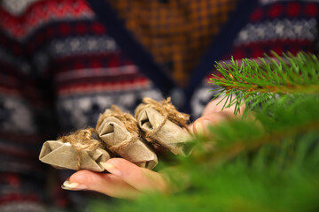 Female hands with a three little craft gifts. Young hipster woman in Christmas sweater holding 3 organic gifts, rope bow. Cozy warm winter detail. Green natural foreground