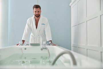 Cheerful man in soft bathrobe leans onto hydro massage tub with hot water in spa salon