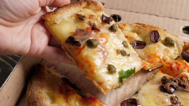 Taking one piece of Fresh baked italian pizza Alla Putanesca in cardboard box on wooden table