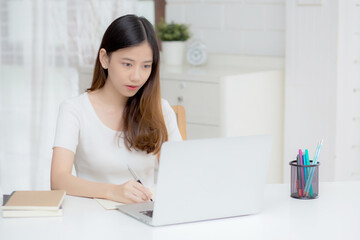 Young asian woman writing on notebook for planning working and using laptop computer on desk at home, girl notes about finance, female study and learning, business and communication concept.