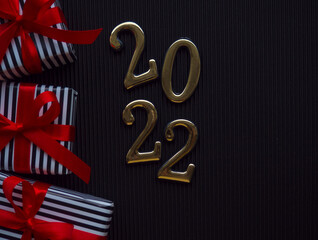 2022 on black background. 2022 gold figures lie near gifts in a stripped black and white wrapper and with red ribbons on the dark corrugated cardboard. Christmas and New year card with place for text