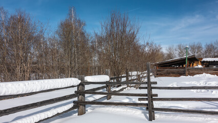 Thick layers of snow lie on an old unpainted wooden fence. Snowdrifts on the ground. Birch grove against the blue sky. Outbuildings are visible in the distance. Siberia.  Taltsy