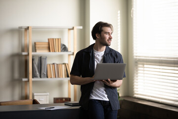 Dreamy handsome young businessman with computer in hands looking in distance, enjoying break pause time in modern home office, planning new project, visualizing successful future, leaning on table.