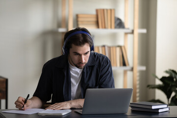 Happy young man in wireless headphones watching educational lecture on computer, enjoying studying on online courses, holding video call with mentor improving knowledge writing notes in copybook.