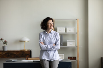 Happy dreamy beautiful middle aged older woman looking away, standing with folded arms in modern...