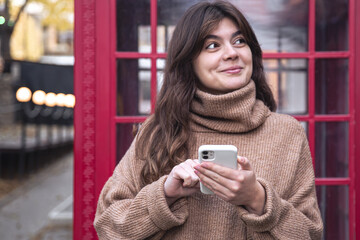 Cute young woman with a smartphone on the background of a red telephone booth.