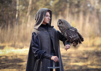 Male warrior in grey cloak and long sword in hand with owl on his shoulder standing in the forest. Fantasy portrait of a youn man in warrior costume - obrazy, fototapety, plakaty