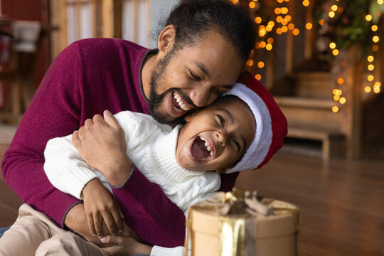 Overjoyed young African American father and small son have fun play together on Christmas at home. Happy biracial dad and little boy child feel playful laugh tickle enjoy New Year winter holidays.