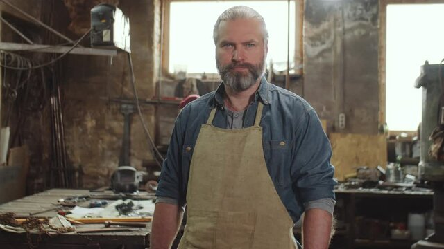 Portrait of bearded blacksmith in apron standing in smithy and looking at camera with confidence
