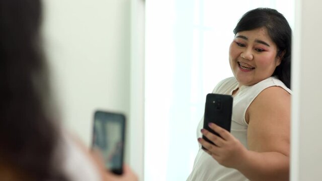 Body positive plus size female clothing and holding mobile phone taking selfie photo using smartphone camera. Body positive plus size woman happily and is proud of herself looking at mirror at home